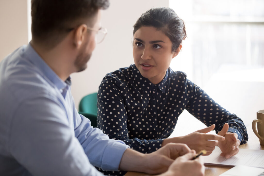 Young female mentor coach worker talking to male coworker teaching intern having business conversation with workmate, serious hindu manager helping colleague discussing new project in office