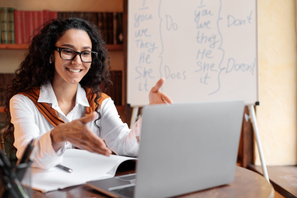 Online Education. Female English teacher having video conference chat with students and class group, using laptop. Woman wearing glasses and wireless earphones, talking to webcam, explaining