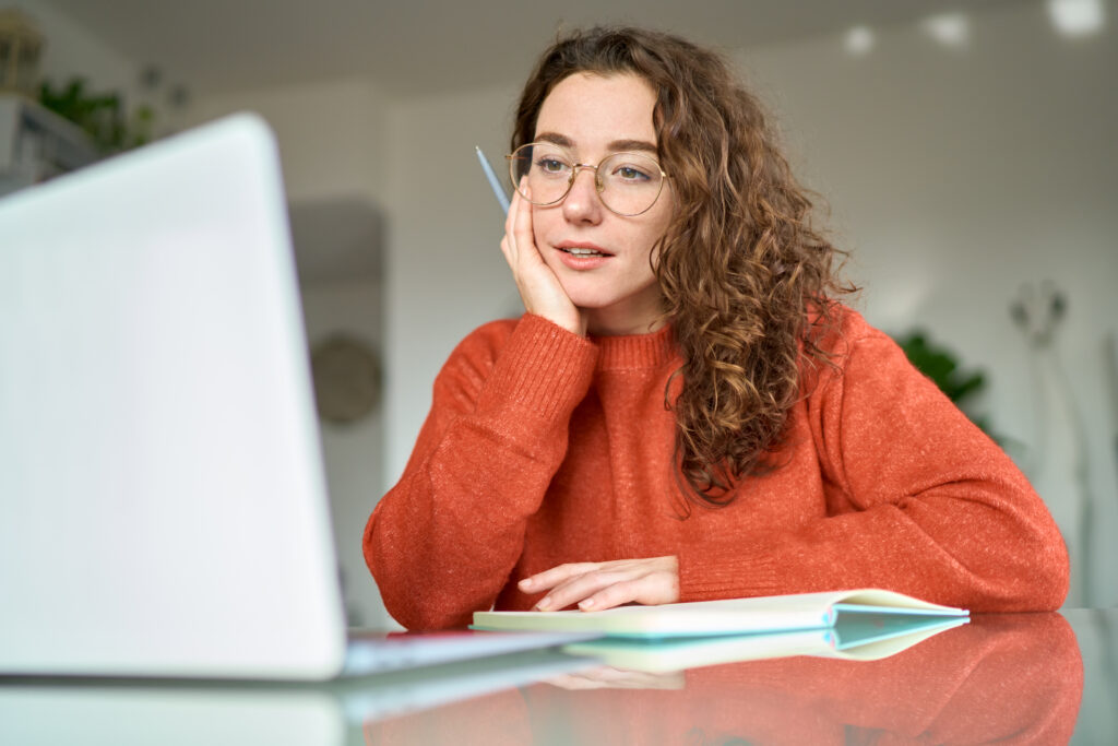 Young female student sitting infront of her laptop with a pen in her hand and a notebook, attentively listening to the teacher