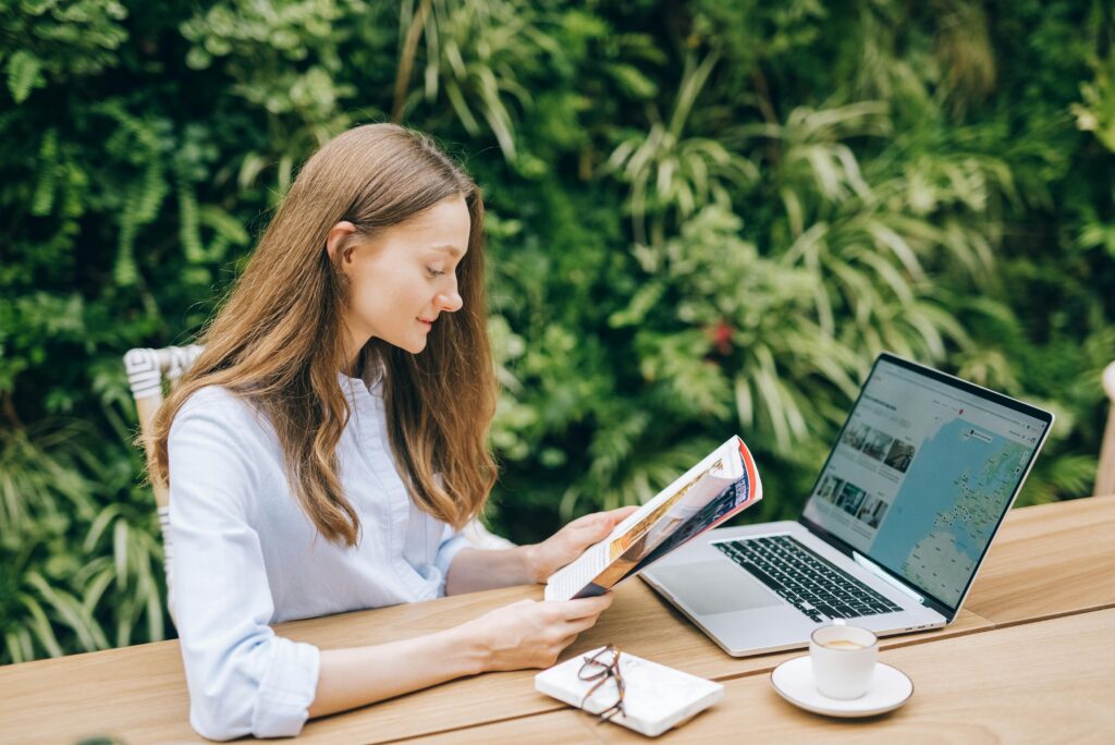 Frau sitzt im Garten und informiert sich über eine Reise, indem sie an Ihrem Laptop die Karten checkt und nebenbei ein Magazin liest