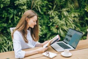 Frau sitzt im Garten und informiert sich über eine Reise, indem sie an Ihrem Laptop die Karten checkt und nebenbei ein Magazin liest