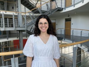 Mays Zatar, Praktikantin bei SprachUnion. Sie trägt ein weißes Kleid, eine Brille hat dunkles, gelocktes Haar und steht im Treppenhaus des Bürogebäudes von SprachUnion.