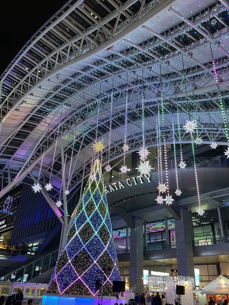 Ein modernes Glasgebäude in Japan ist mit Leuchtsternen behangen, die von dem Dach herunterhängen. Vor dem Gebäude steht ein Tannenbaum mit einem leuchtenden Stern auf der Spitze. Er ist umwunden von bunt leuchtenden Lichterketten.