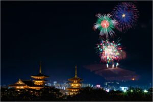Es ist Nacht. Im Vordergrund steht ein eleuchteter, japanischer Tempel. Im Hintergrund sieht man leicht die Konturen von modernen Gebäuden. Der Himmel ist erstrahlt von buntem Feuerwerk.