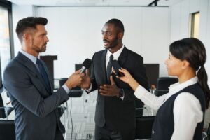 Im Bild sind zwei Männer und eine Frau in Anzügen zu erkennen, welche sich in einem eintönigen Büroraum befinden. Im Vordergrund stehen die drei Personen, wobei der Mann in der Mitte den anderen Beiden ein Interview gibt. Die Frau rechts hält ein Handy zur Tonaufnahme in die Höhe und der andere Mann links ein Mikrofon. Dabei ist die Mimik der Beteiligten neutral. Im Hintergrund sind leere Stühle und ein Rednerpult abgebildet.